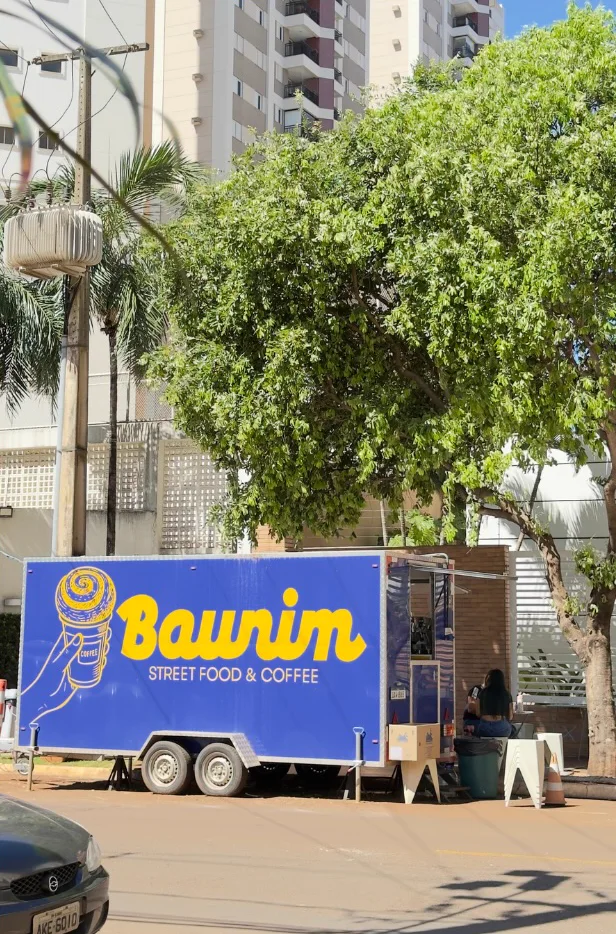 O trailer azul da Baunim estacionado na sombra de uma árvore, com os prédios da Gleba Palhano ao fundo